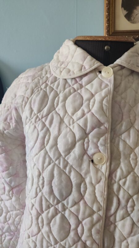 Penhoar vintage em matelassê claro com botões frontais, apresentando desgaste no tecido acolchoado e marcas do tempo, exibido em manequim.