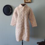 Penhoar vintage em matelassê claro com botões frontais, apresentando desgaste no tecido acolchoado e marcas do tempo, exibido em manequim.