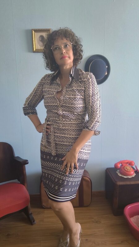 Vestido chemisier vintage original dos anos 70 na cor branca com estampa geométrica azul marinho. Modelo midi com mangas longas, fechamento frontal por botões e faixa para amarração na cintura. Peça vintage feminina em excelente estado de conservação, fotografada em ambiente interno com decoração retrô.