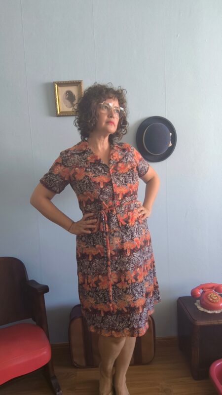 Chemisier vintage original dos anos 70 em jersey de poliamida com estampa floral em tons coral, marrom e bege. Modelo midi com mangas curtas, fechamento frontal por botões e faixa para amarração na cintura. Peça feminina vintage em excelente estado de conservação, fotografada em ambiente interno com decoração retrô.