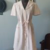 Penhoar vintage em piquê branco com bordados florais, bolso frontal e faixa rosa de cetim para ajuste da cintura.