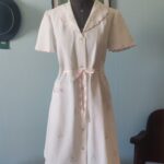 Penhoar vintage em piquê branco com bordados florais, bolso frontal e faixa rosa de cetim para ajuste da cintura.