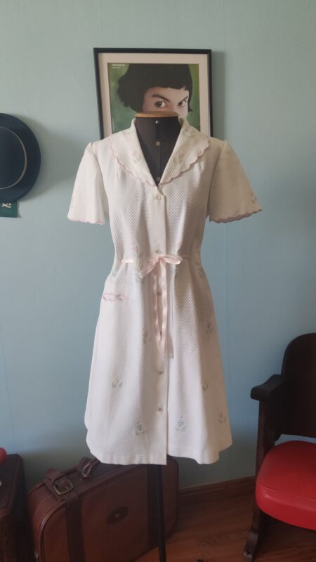 Penhoar vintage em piquê branco com bordados florais, bolso frontal e faixa rosa de cetim para ajuste da cintura.