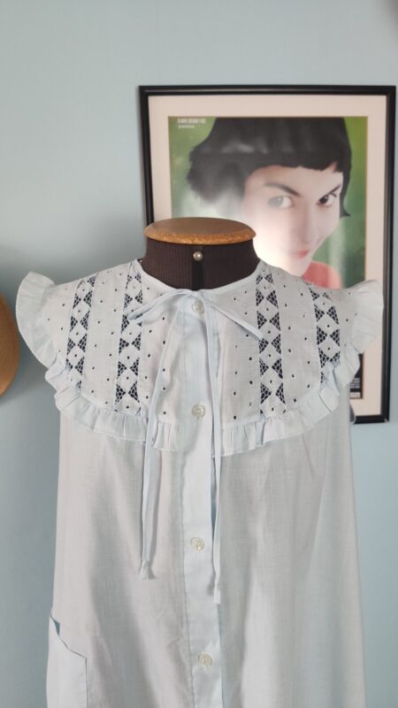 Close da parte superior de camisola vintage azul-claro mostrando a pala com bordado inglês, babados, laço frontal e pequenos botões.