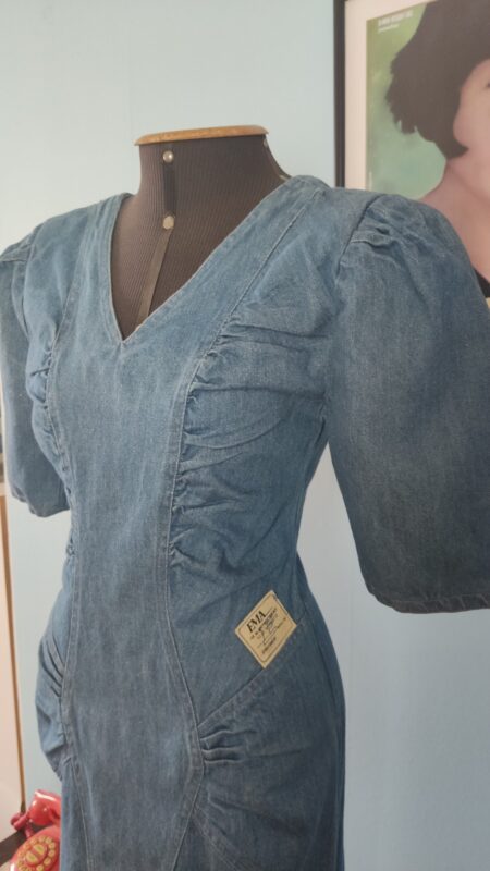 Vestido jeans vintage original dos anos 80 em manequim, com ombreiras, franzidos laterais e silhueta ajustada.